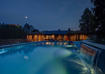 A pool with lights on and a house in the background.